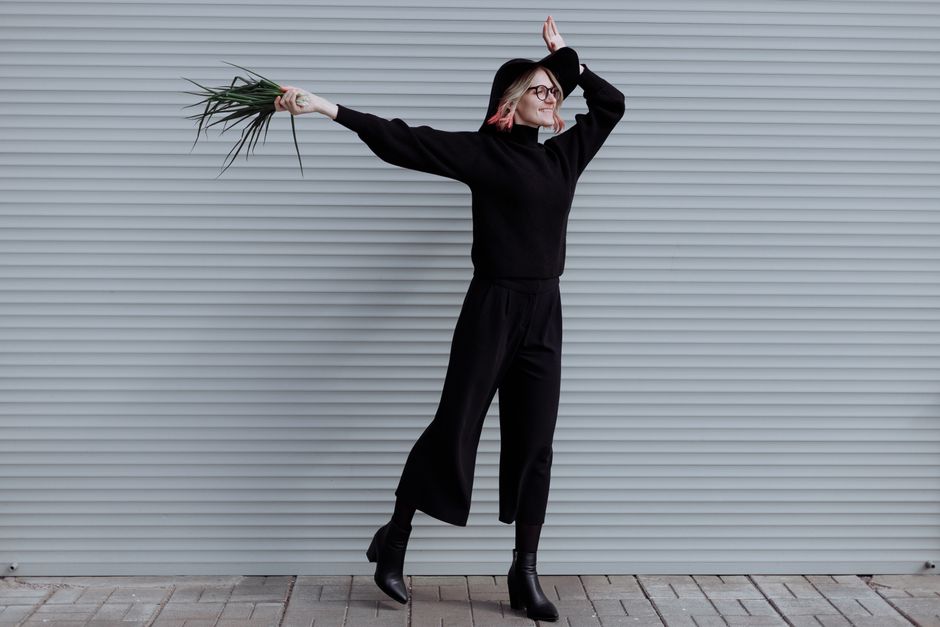 A woman in black jumps energetically holding green leaves against a corrugated backdrop.