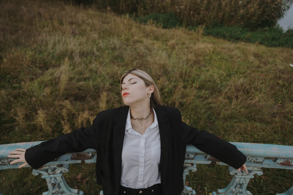 Elegant young woman posing outdoors in a field with eyes closed, exuding fashion and grace.