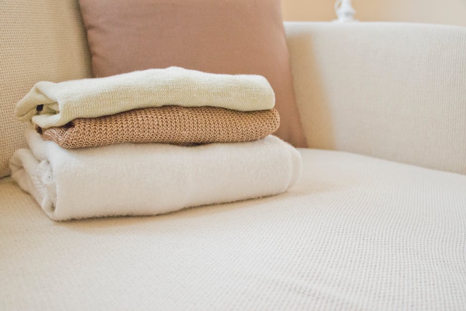 Soft folded sweaters in warm tones on a cozy cream sofa, showcasing home comfort and style.