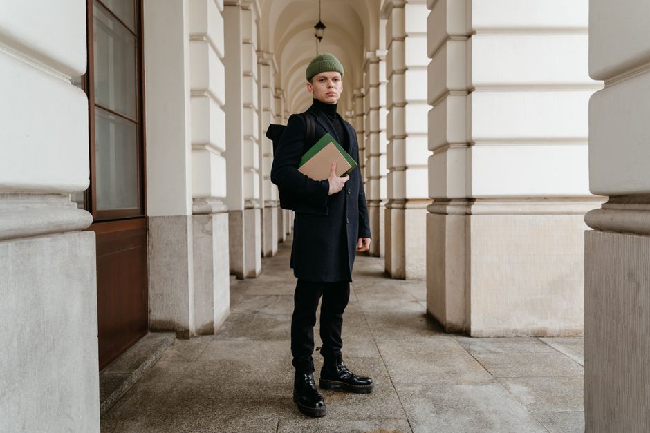A young man in a dark coat and beanie stands in a classic archway holding books.