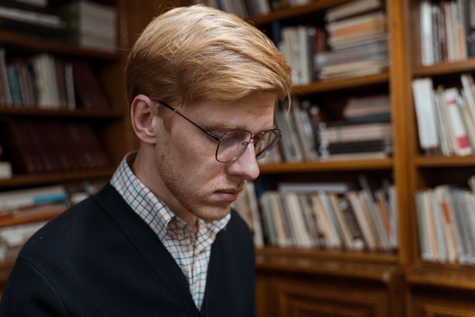 A young man with glasses focused on reading in a library environment, surrounded by books.