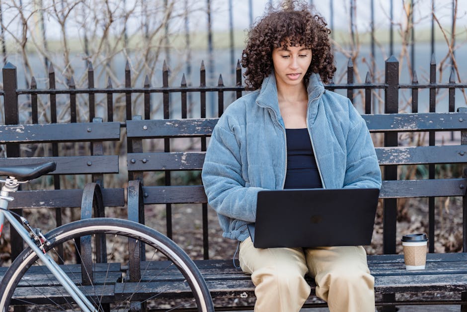 Focused young ethnic woman with curly brown hair in stylish warm jacket sitting on wooden bench in city park and typing on laptop during remote work