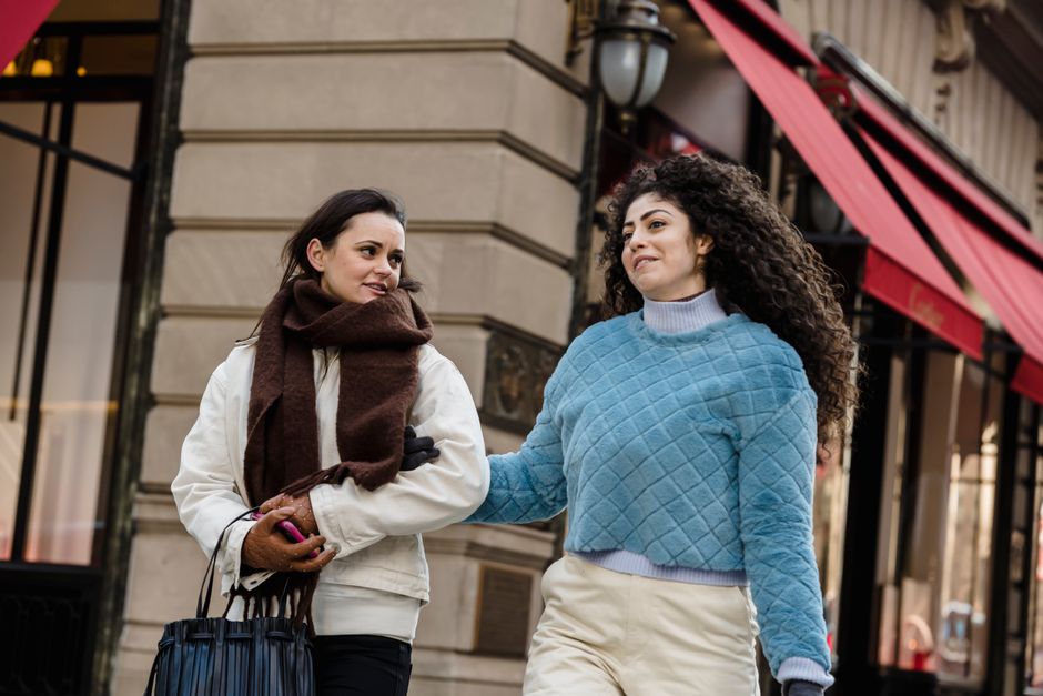 Low angle of stylish young diverse ladies in warm clothes holding hands while walking on city street near modern boutiques and communicating