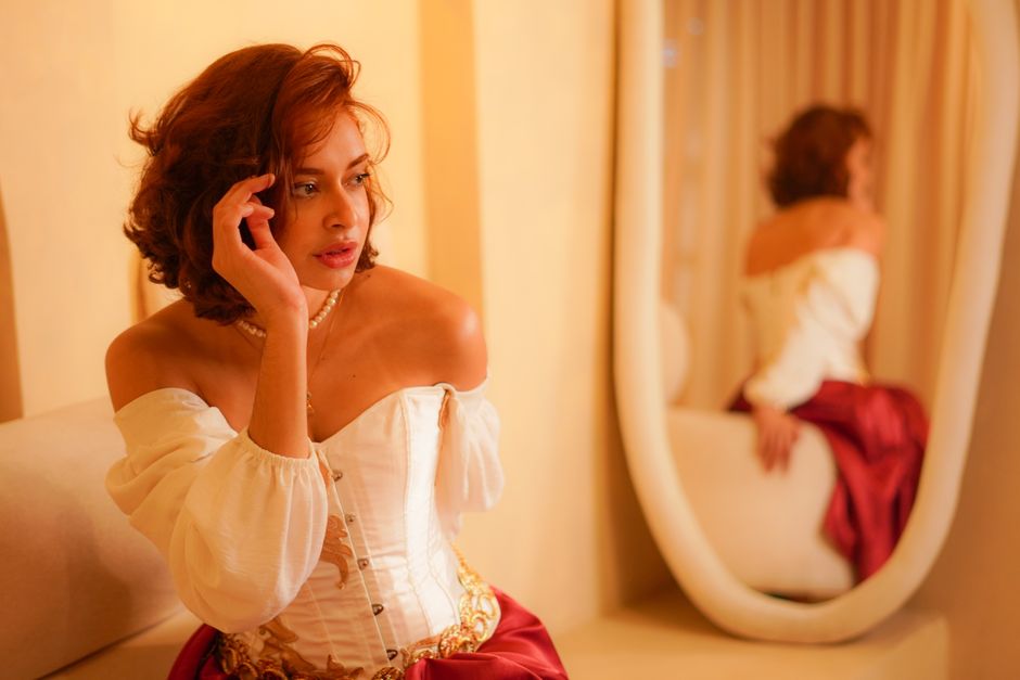 A woman in a vintage outfit with a corset and red skirt, seated indoors with a mirror reflection.