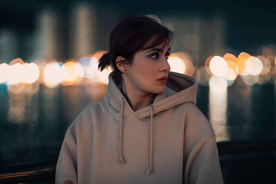 A thoughtful woman in a hoodie gazes into the distance amid colorful night lights.