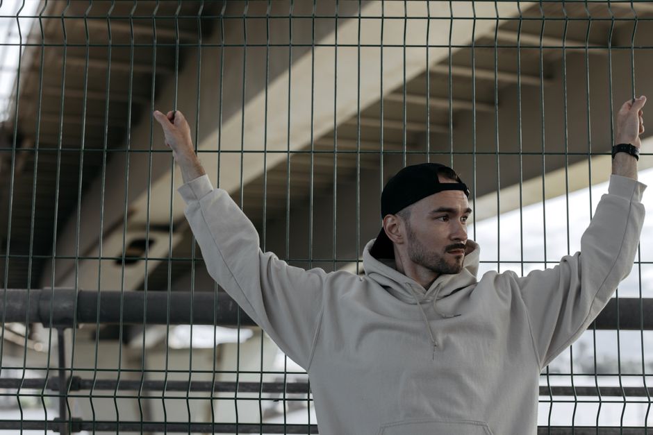 Young man in casual hoodie and cap by a chain link fence, outdoors under a bridge.