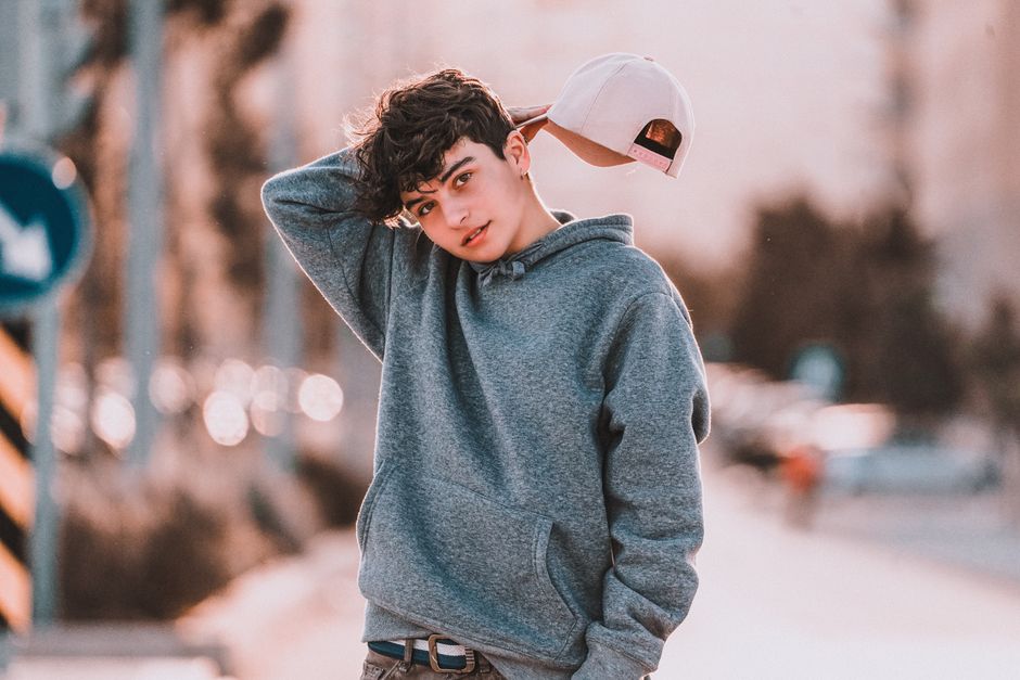Contemplative teenager in stylish apparel with raised arm standing with tilted head on street while looking at camera