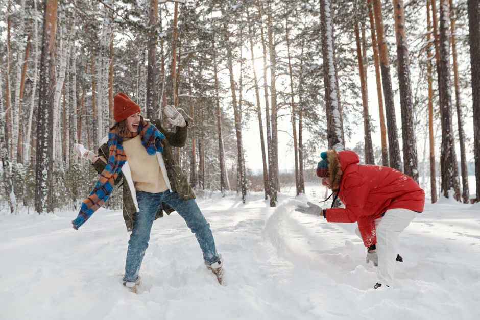 Two adults playfully enjoying a snowball fight in a serene winter forest setting.