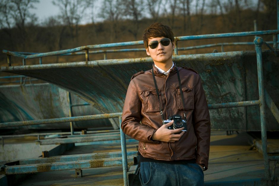 A man in sunglasses and jacket holds a camera in an outdoor industrial setting.