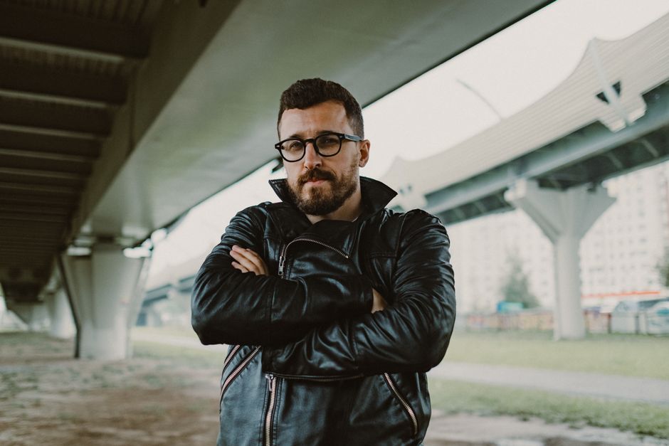 Man with glasses and beard posing under a city bridge in a leather jacket.