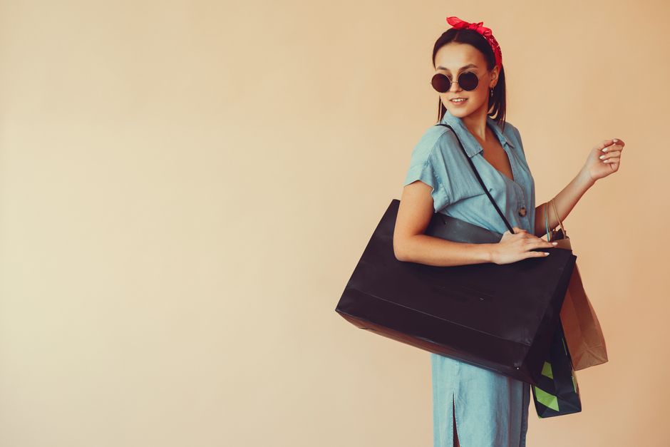 Side view of happy young shopaholic female wearing trendy blue dress and sunglasses carrying many shopping bags in studio on beige background