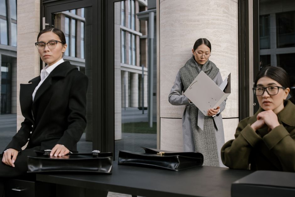 Three professional women in formal outfits with briefcases, posing outside a modern office building.