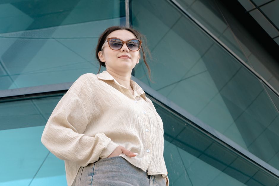 Confident woman wearing sunglasses poses against modern glass architecture backdrop.