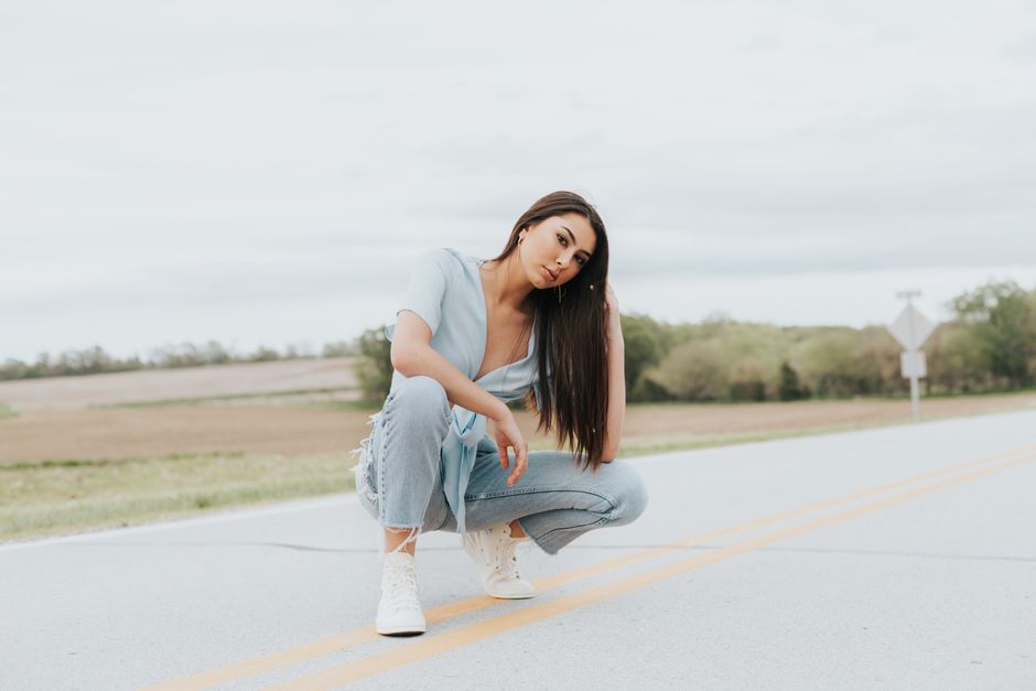 Asian woman kneeling stylishly on a rural road wearing denim and white shoes.