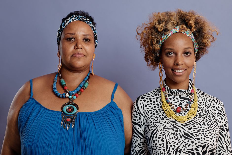 Vibrant portrait of two women in stylish outfits and bold jewelry against a purple background.