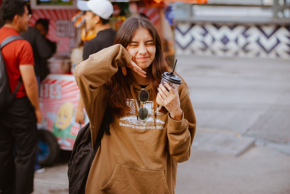 A young woman with a drink, squinting happily in a street market setting.