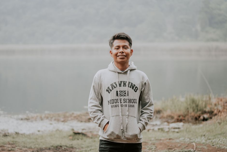Casual portrait of a smiling young man in a hoodie standing outdoors by a misty lake.