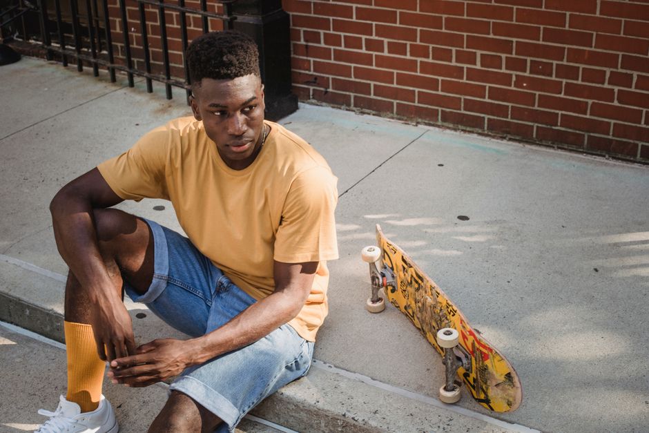 From above of stylish young African American male millennial in yellow t shirts and shorts relaxing on sidewalk near skateboard and looking away after riding