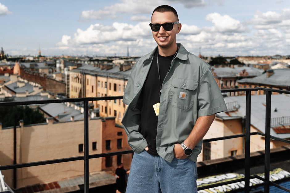 Smiling man with sunglasses on a rooftop with cityscape in the background under a bright sky.