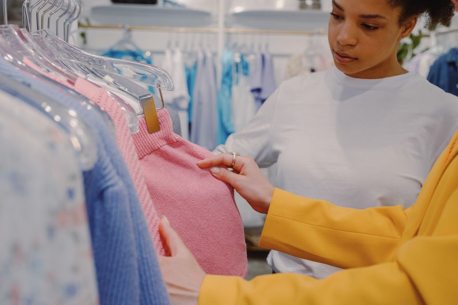Young African American female customer with friend choosing pink sweater in range of knitted clothes in shop